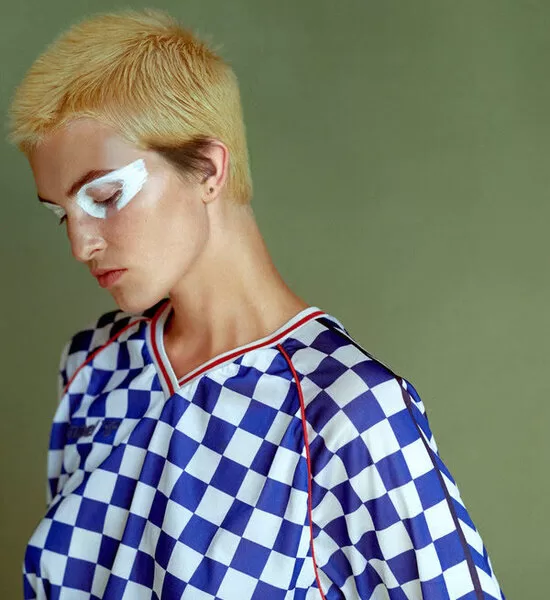 Fotografie v galerii od Jan Balcařík, který vytvořil vzhled této ženy. Zelené pozadí, žena odbarvený blond na dost krátké vlasy. Fotka je z profilu na její levou stranu. Na sobě má kostkované modrobílé tričko.